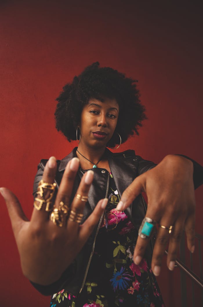 Stylish woman with afro showcasing bold rings and jewelry against a red background.